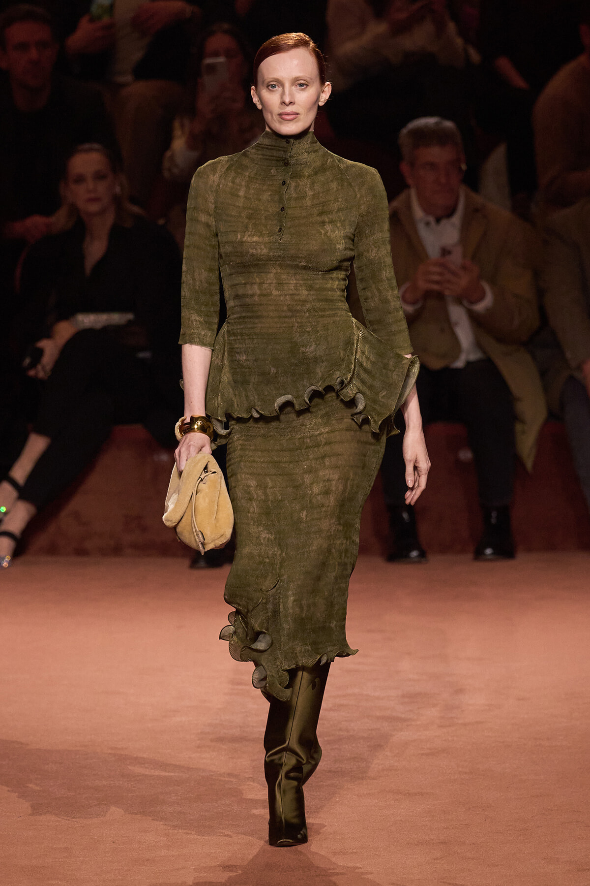Model walks the runway in an olive green, textured dress with a high neckline and ruffled peplum, carrying a tan handbag; audience in dim light behind.