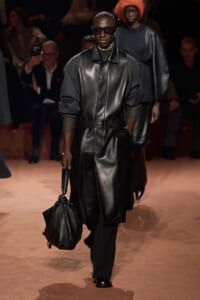Fashion model walking a runway in a black leather coat with a belt, dark pants, and large sunglasses, carrying a black bag.