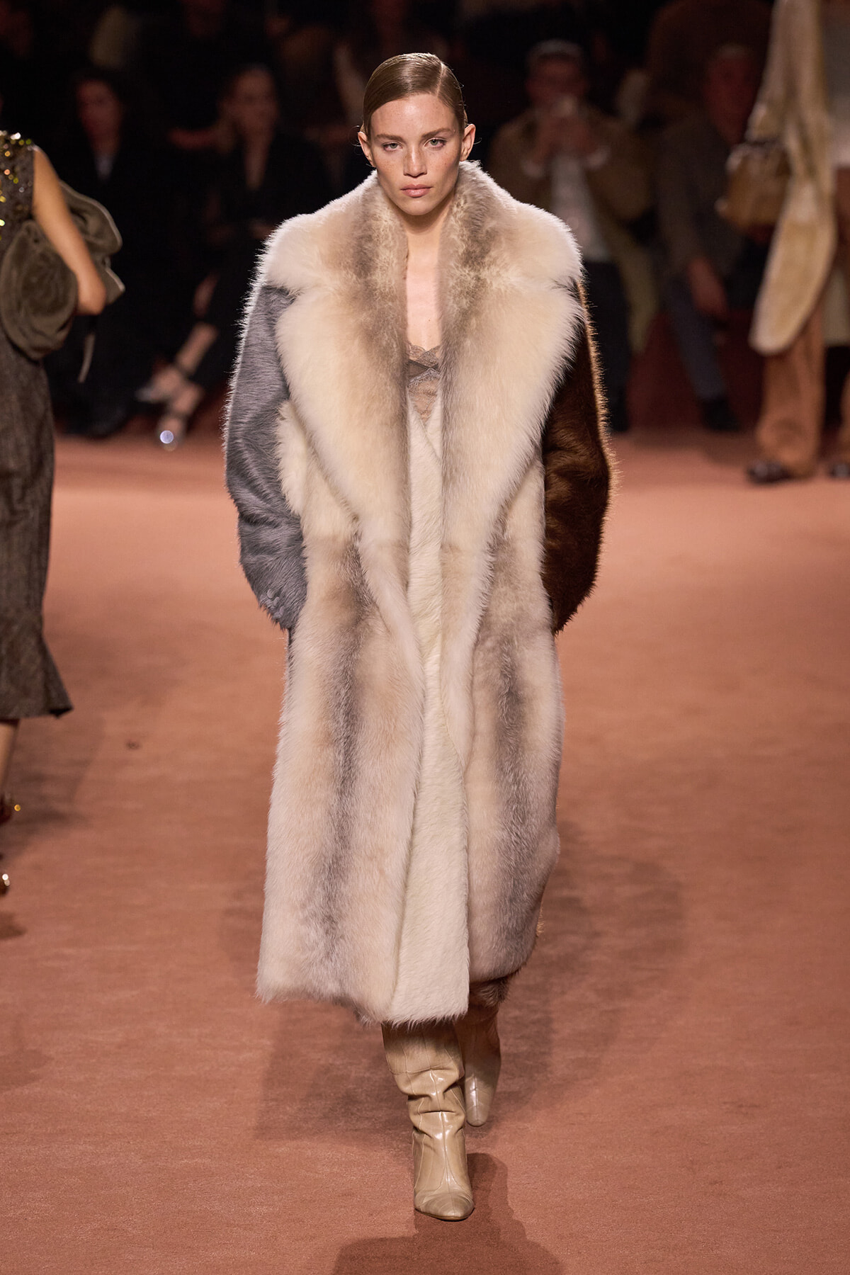 Model walking a fashion runway in a long, color-block fur coat and cream boots.