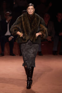 Model walks a fashion runway wearing a dark fur coat over a textured gray midi skirt and shiny black knee-high boots; audience sits in the background.