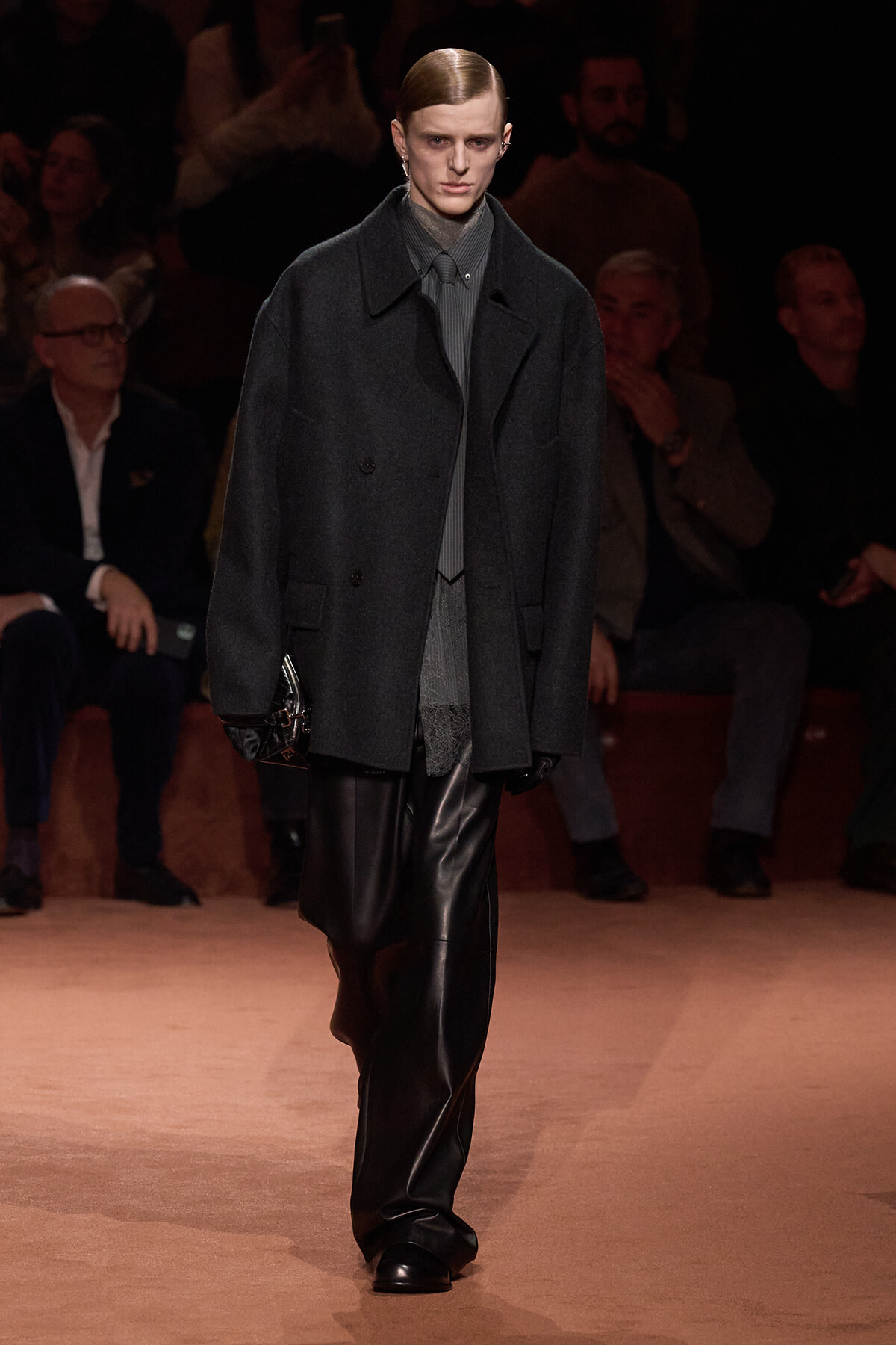 Male model walks a fashion runway in a dark oversized coat, gray shirt, and black leather pants, holding a small clutch; audience in dim background.
