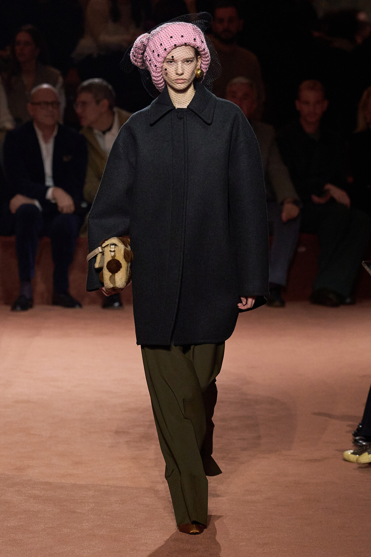 Runway model wearing a large dark coat, olive pants, and a pink netted hat, carrying a small fur-trimmed handbag.