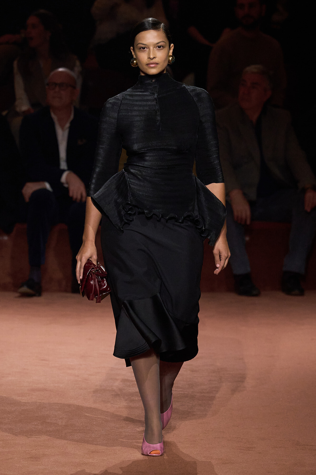 Model on a runway wearing a black peplum top with a high neck and three-quarter sleeves, a black skirt, pink open-toe heels, and a burgundy clutch.
