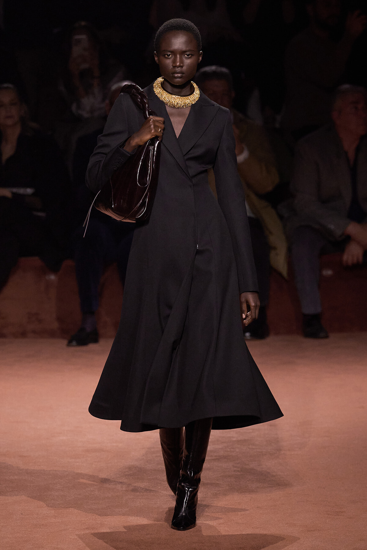 Model walks a runway in a black knee‑length coat dress, with a bold gold necklace, black boots, and a large glossy brown handbag.