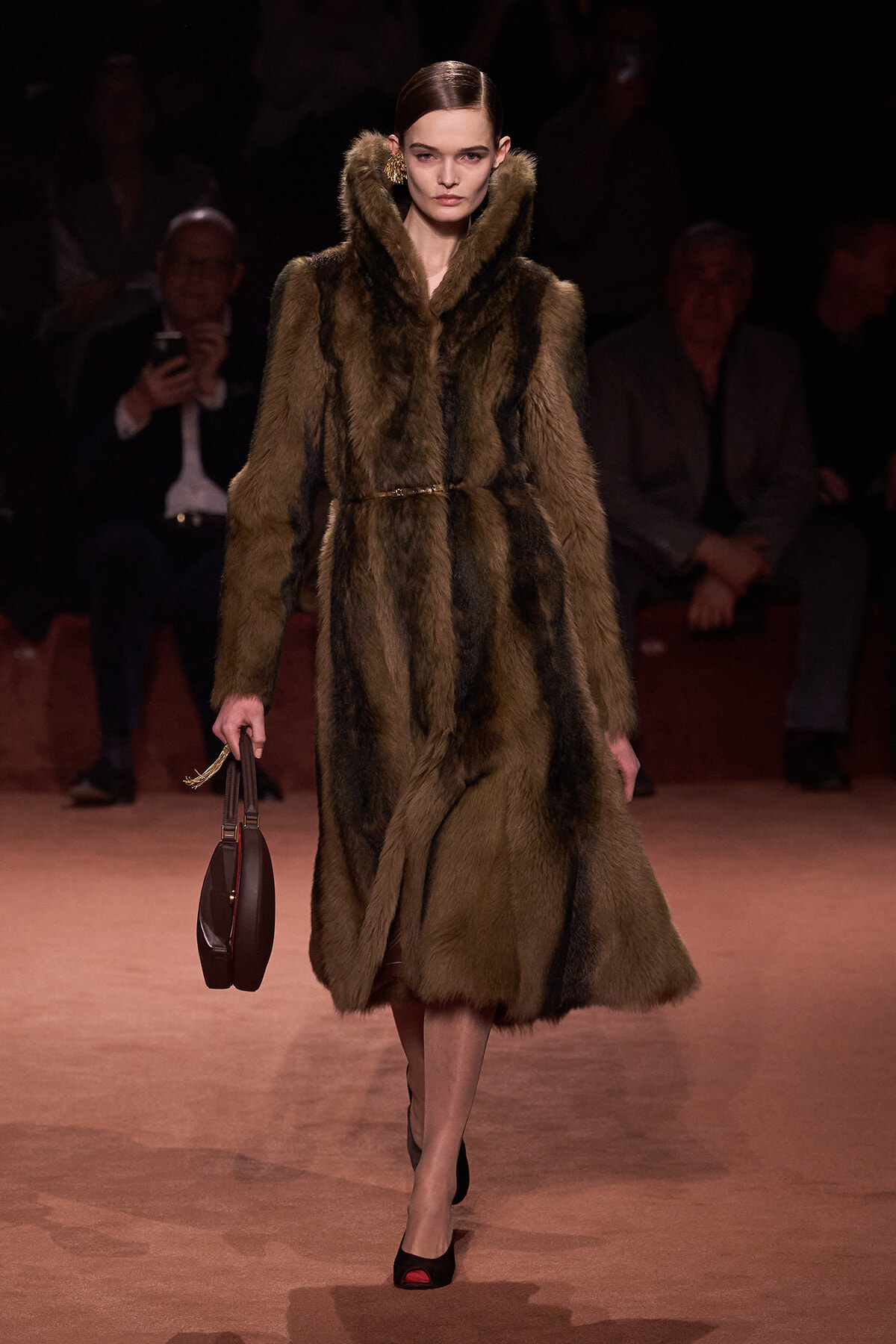 Model walks the runway in a long brown fur coat with a belt, carrying a dark rounded handbag; audience in dim background.