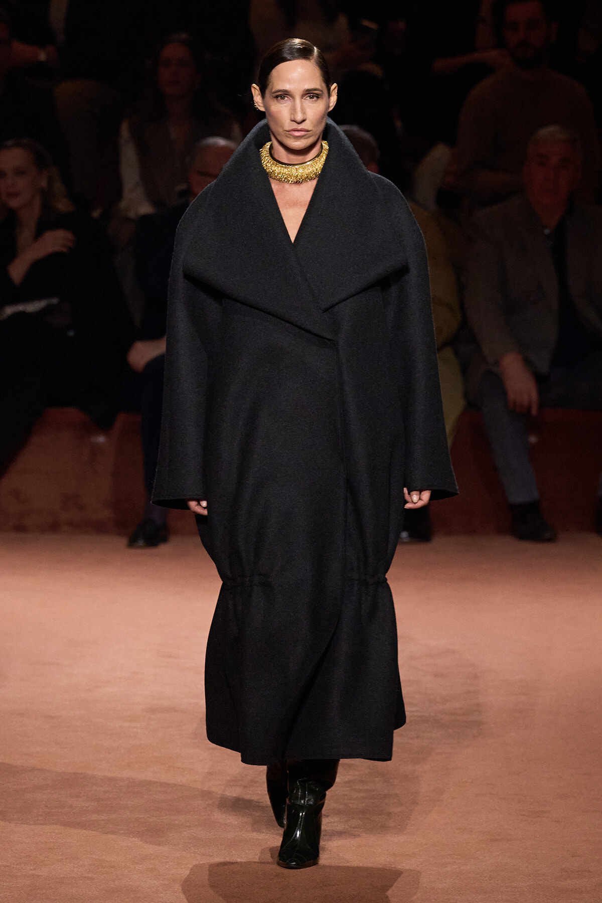 Model walks the runway in an oversized black coat with a wide lapel, paired with a chunky gold necklace and dark boots.