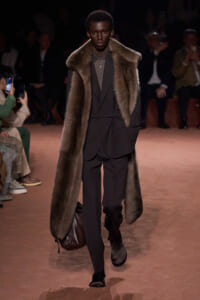 Male model walks the runway in a dark suit with an oversized fur coat draped over the shoulders, carrying a brown leather bag.