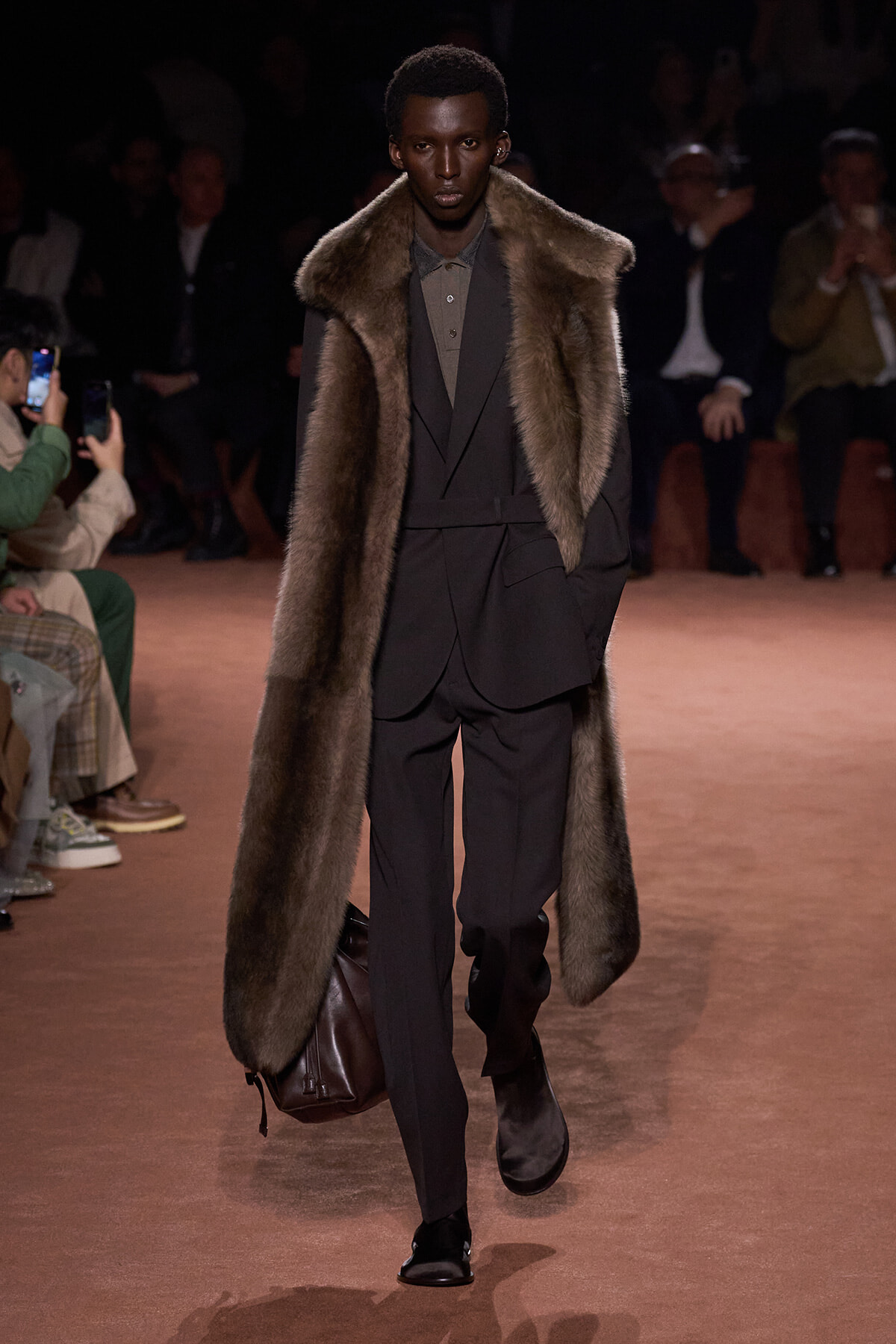 Male model walks the runway in a dark suit with an oversized fur coat draped over the shoulders, carrying a brown leather bag.