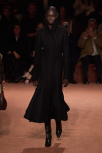 Model walks the runway in a long black hooded coat and matching boots, her expression neutral as lights focus on her.