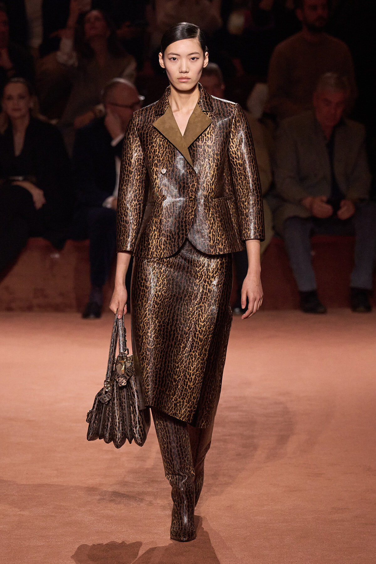 Model walks the runway in a brown leopard-print two-piece suit with matching skirt and a metallic handbag in hand.