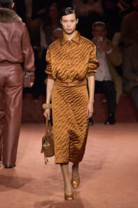 Model walks the runway in a matching bronze quilted two-piece outfit with a high-neck bomber jacket and midi skirt, carrying a brown handbag and wearing tan peep-toe heels.