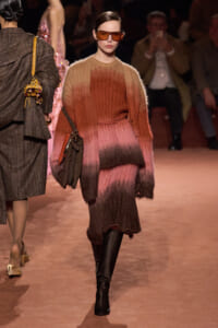 Model walks a runway in a gradient fuzzy knit dress from pink to brown, with orange-tinted sunglasses and a brown crossbody bag paired with black knee-high boots.