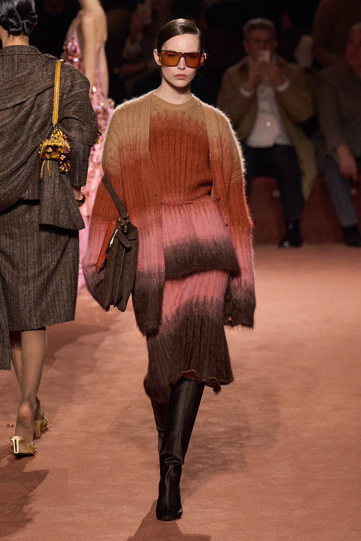 Model walks a runway in a gradient fuzzy knit dress from pink to brown, with orange-tinted sunglasses and a brown crossbody bag paired with black knee-high boots.