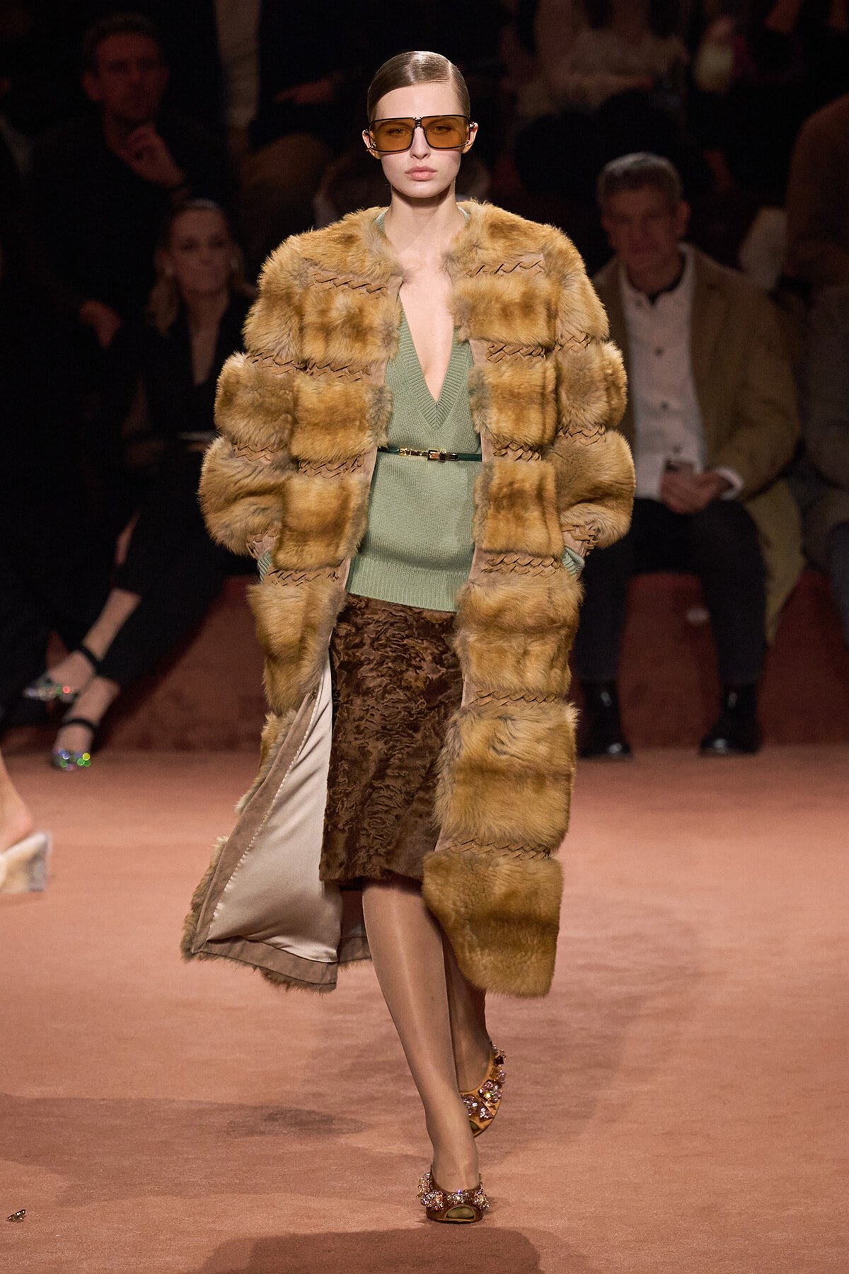 Model strides down the runway in a tan fur-coat with a sage green V-neck sweater and a patterned midi skirt, accessorized with a thin belt and oversized brown sunglasses.