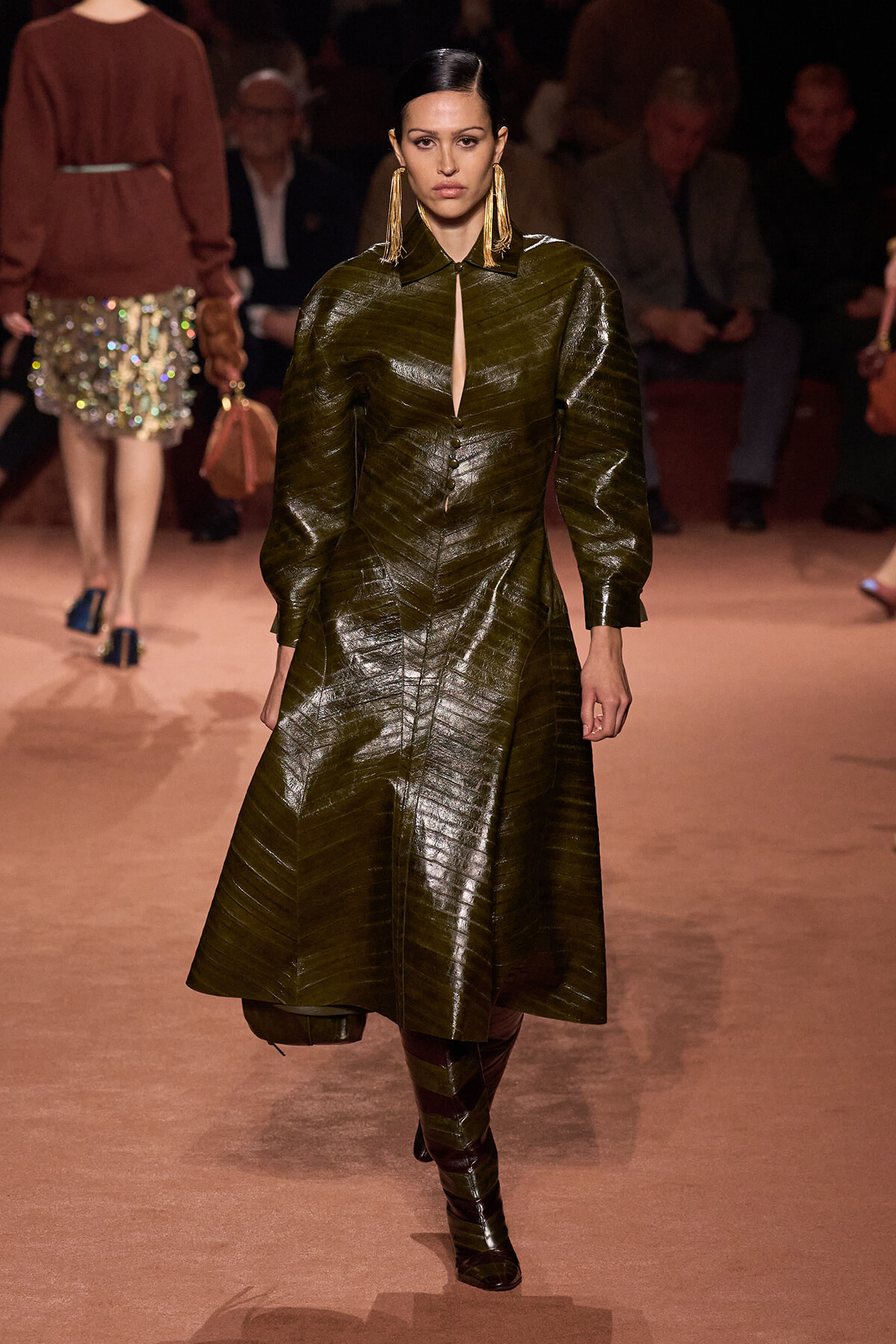 Model walking the runway in a dark olive-green, metallic trench-style coat with structured, voluminous sleeves and a front slit, paired with tall boots and statement earrings.