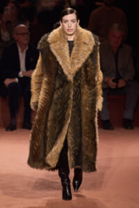 Model walking the runway in a long brown fur coat with large lapels, black outfit, and knee-high boots.
