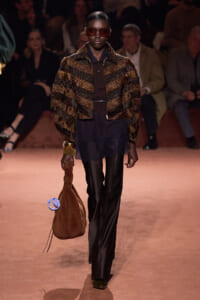 Model walks the runway in a dark outfit with a brown striped fur jacket, oversized sunglasses, and black wide-leg pants, carrying a brown suede bag with a blue circular accessory.