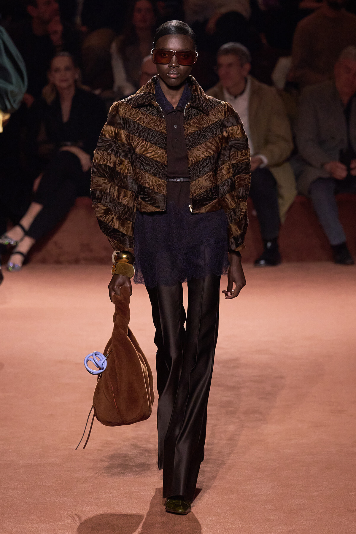Model walks the runway in a dark outfit with a brown striped fur jacket, oversized sunglasses, and black wide-leg pants, carrying a brown suede bag with a blue circular accessory.