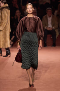 Model walks the runway in a burgundy satin blouse with voluminous sleeves and a dark emerald quilted midi skirt, carrying a burgundy handbag.