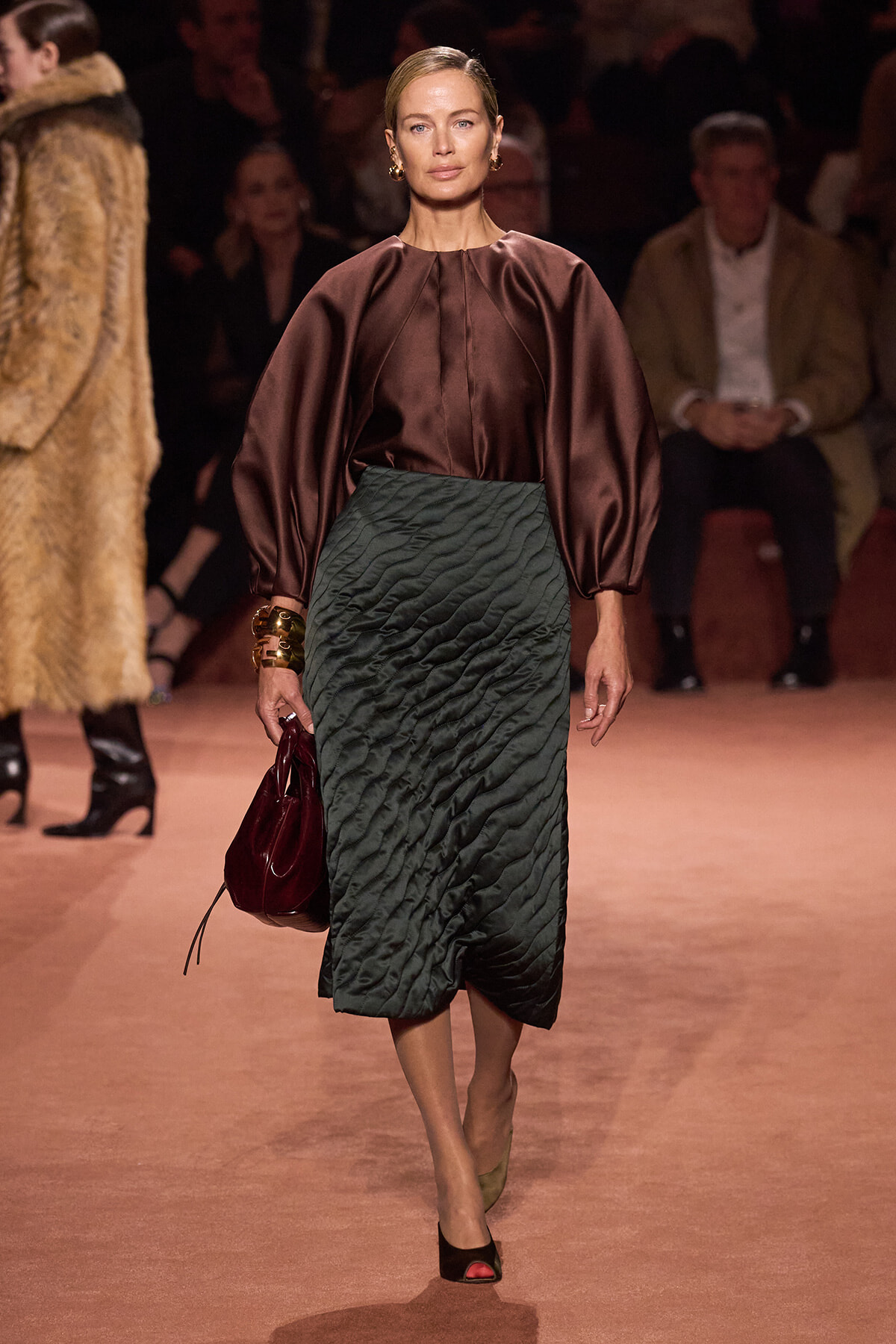 Model walks the runway in a burgundy satin blouse with voluminous sleeves and a dark emerald quilted midi skirt, carrying a burgundy handbag.
