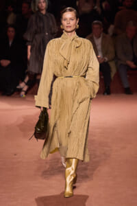 Model walking a runway in a camel-colored pleated midi dress with a neck tie, belt, and gold knee-high boots, holding a dark handbag.