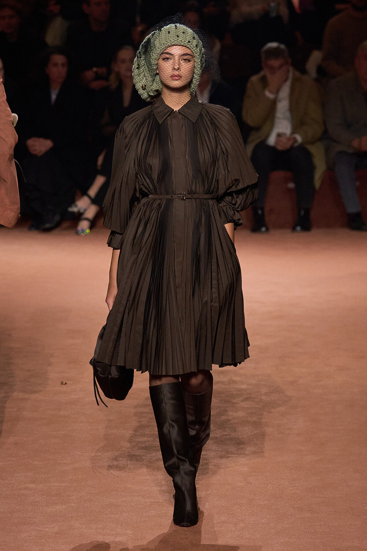 Model walks a runway in a dark brown pleated dress with a belt, tall black boots, and a green knotted headpiece with veil.
