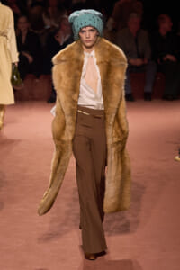 Model walking the runway in a long brown fur coat over a pale striped blouse and brown wide-leg pants, with a teal knit turban.