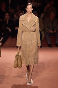 Model walks the runway in a beige perforated coat belted at the waist, carrying a fur handbag and wearing fur-trimmed shoes.