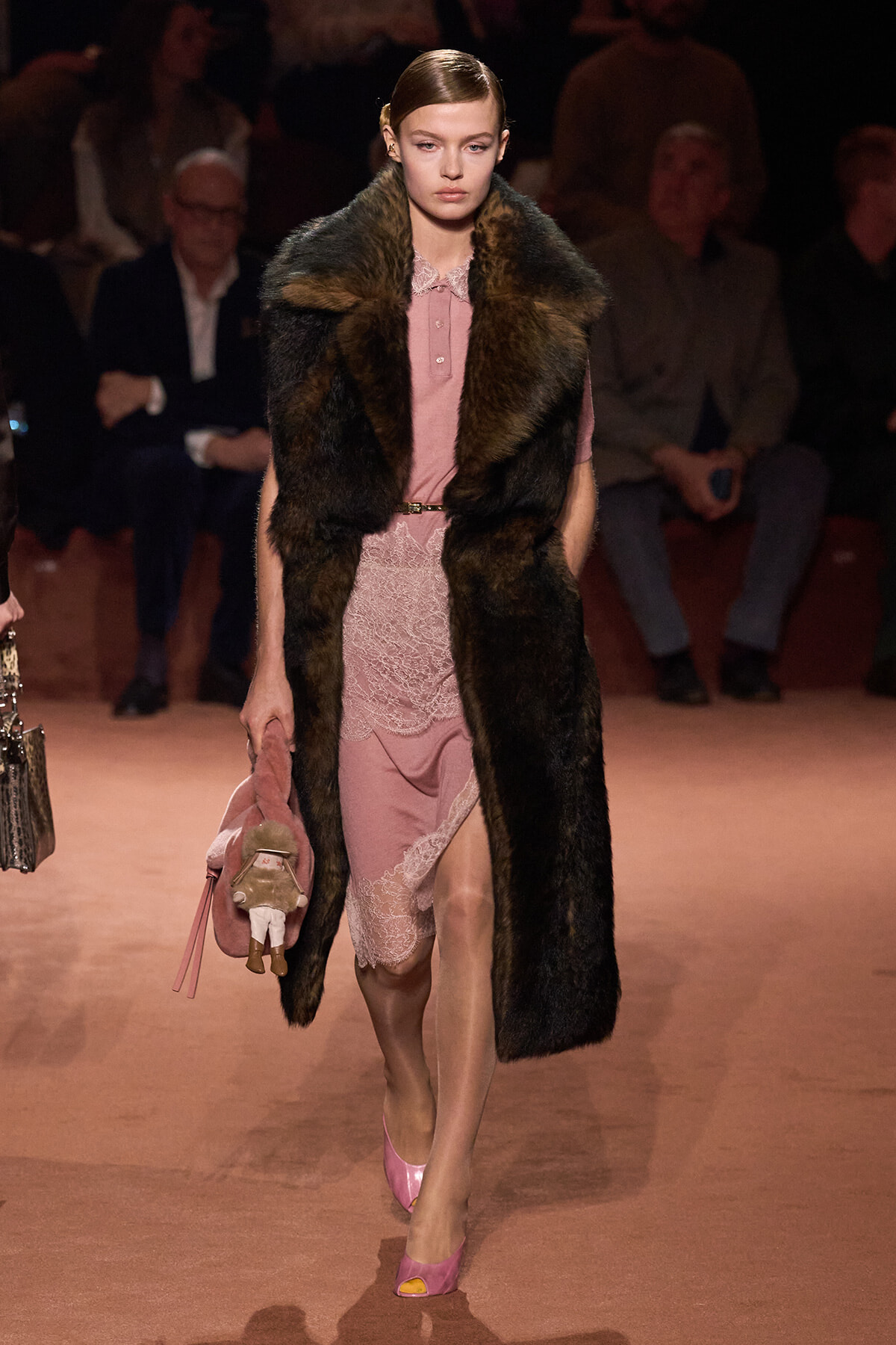 Model walking a runway in a pink lace dress with a dark fur vest and pink heels, carrying a pink coat and small plush accessory.