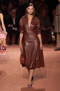 Model walking a runway in a brown leather midi dress with a matching brown handbag and gold earrings