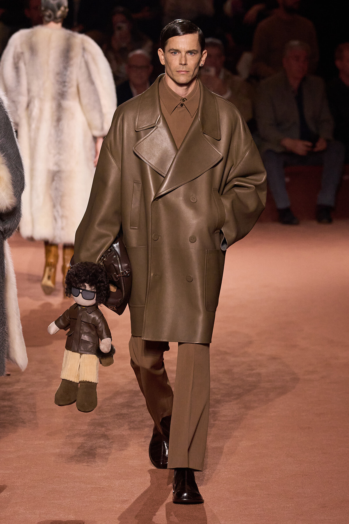 Male model on a runway wearing a large brown leather coat and matching outfit, holding a small plush puppet at his side.