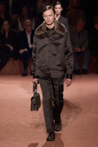 Male fashion model walks the runway in a dark satin suit with a fur collar, carrying a small patterned handbag.