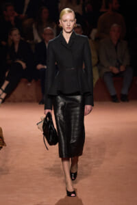 Model walking a fashion runway in a tailored black blazer and black leather midi skirt, carrying a black bag with accessories.