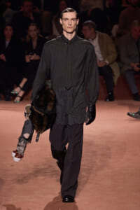 Male fashion model walks the runway in a dark pinstripe shirt and loose black pants, carrying a fur stole and a small child-sized doll hanging by its leg; audience in background