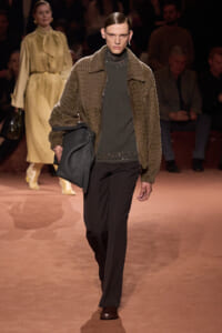 Model on a runway wearing a brown textured jacket over a dark green studded knit sweater, black trousers, brown shoes, and carrying a large black bag.