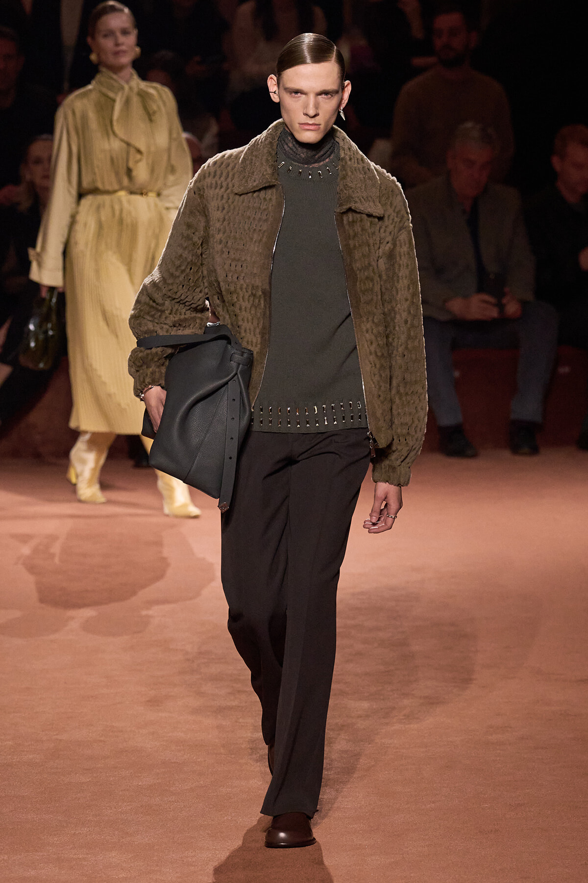 Model on a runway wearing a brown textured jacket over a dark green studded knit sweater, black trousers, brown shoes, and carrying a large black bag.