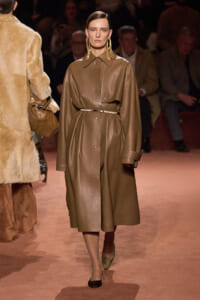 Model walking a fashion runway in a taupe leather trench with a thin belt and oversized sleeves.