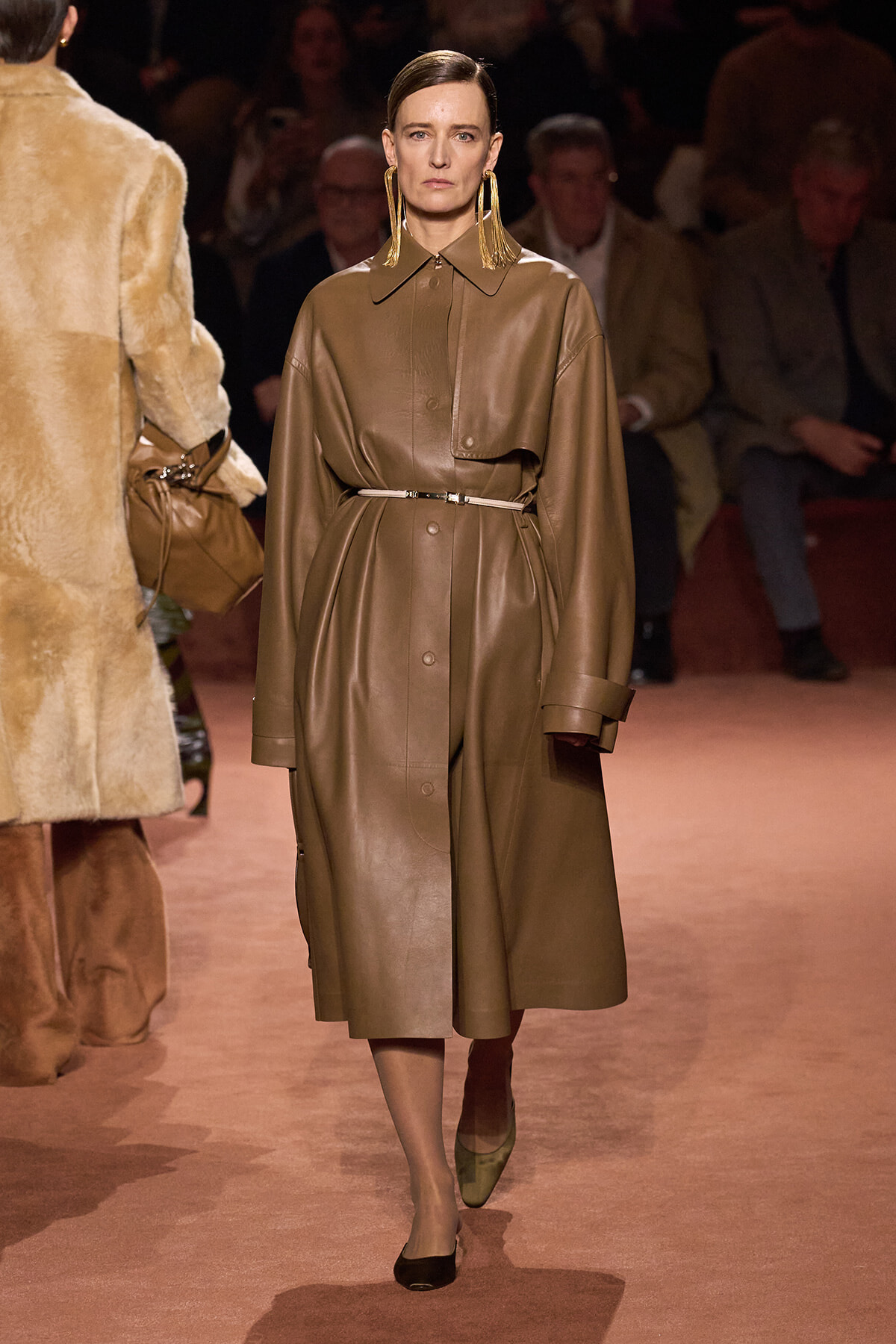 Model walking a fashion runway in a taupe leather trench with a thin belt and oversized sleeves.