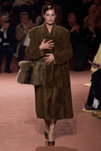 Model on a runway wearing a full-length brown fur coat and holding a matching fur handbag at her side.
