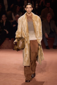 Male model walks a runway in a cream faux-fur coat over a brown gradient knit top and wide brown pants, carrying a tan leather bag on his arm.