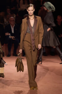 Model walking a runway in a brown tailored suit with burgundy lapels, carrying a brown handbag at her side.