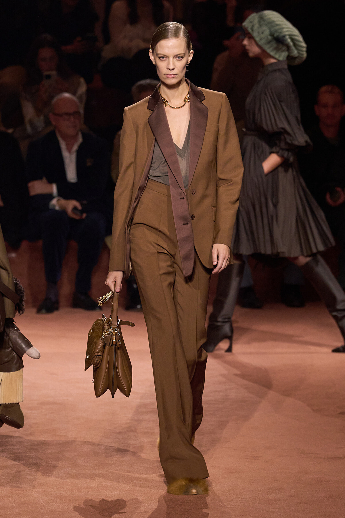 Model walking a runway in a brown tailored suit with burgundy lapels, carrying a brown handbag at her side.