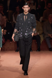 Male runway model wearing a dark gray embellished cardigan over a sheer shirt, black trousers, and a small gray bag; audience in the background.