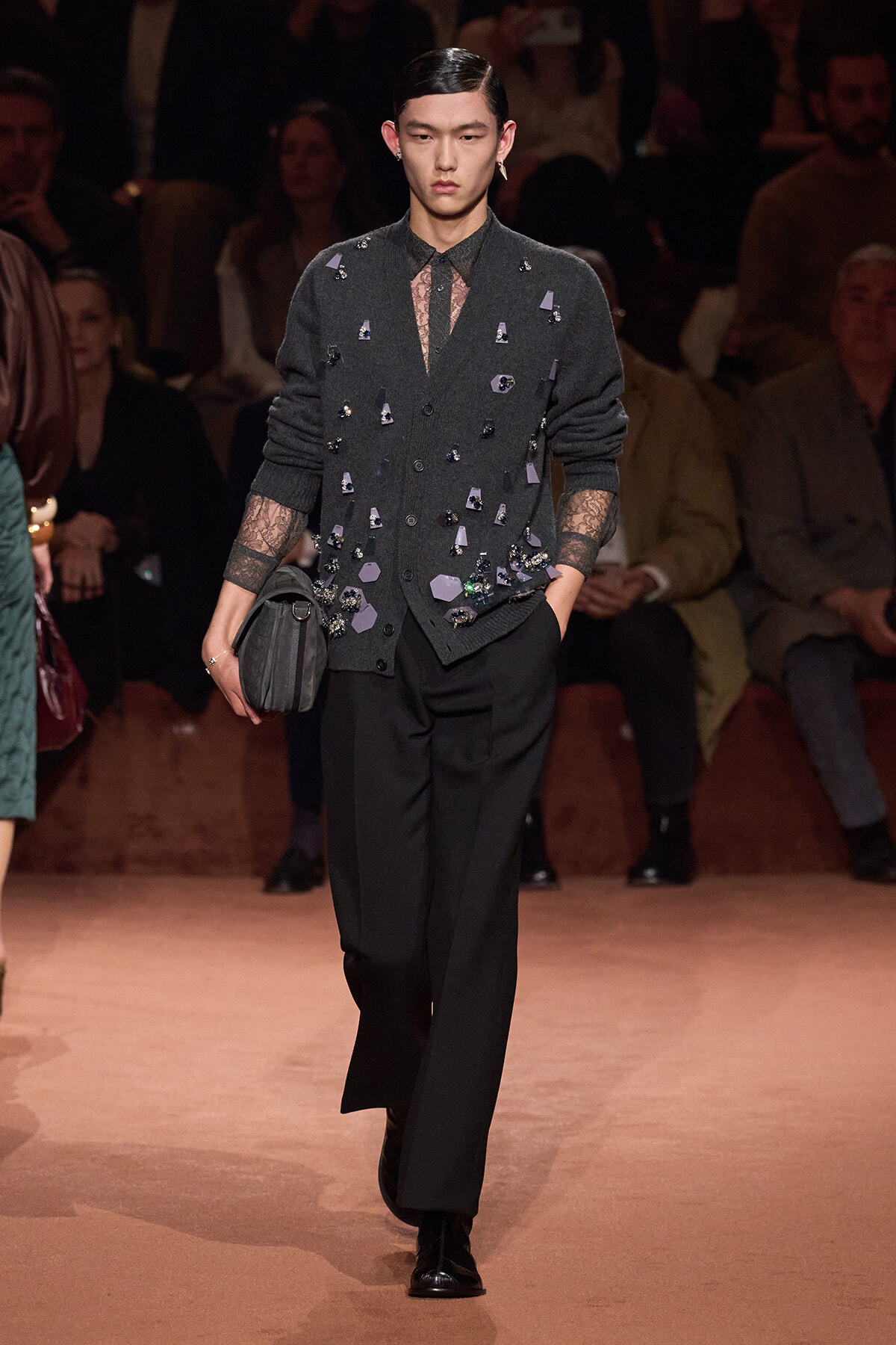 Male runway model wearing a dark gray embellished cardigan over a sheer shirt, black trousers, and a small gray bag; audience in the background.