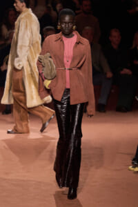 Runway model in rust suede belted coat over pink knit top with shiny black pants and large gold hoop earrings, holding a fur stole far left of center.