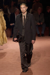 Male runway model in a dark suit with a beige shirt and brown tie, carrying a black clutch as he walks the catwalk. Audience blurred in the background.