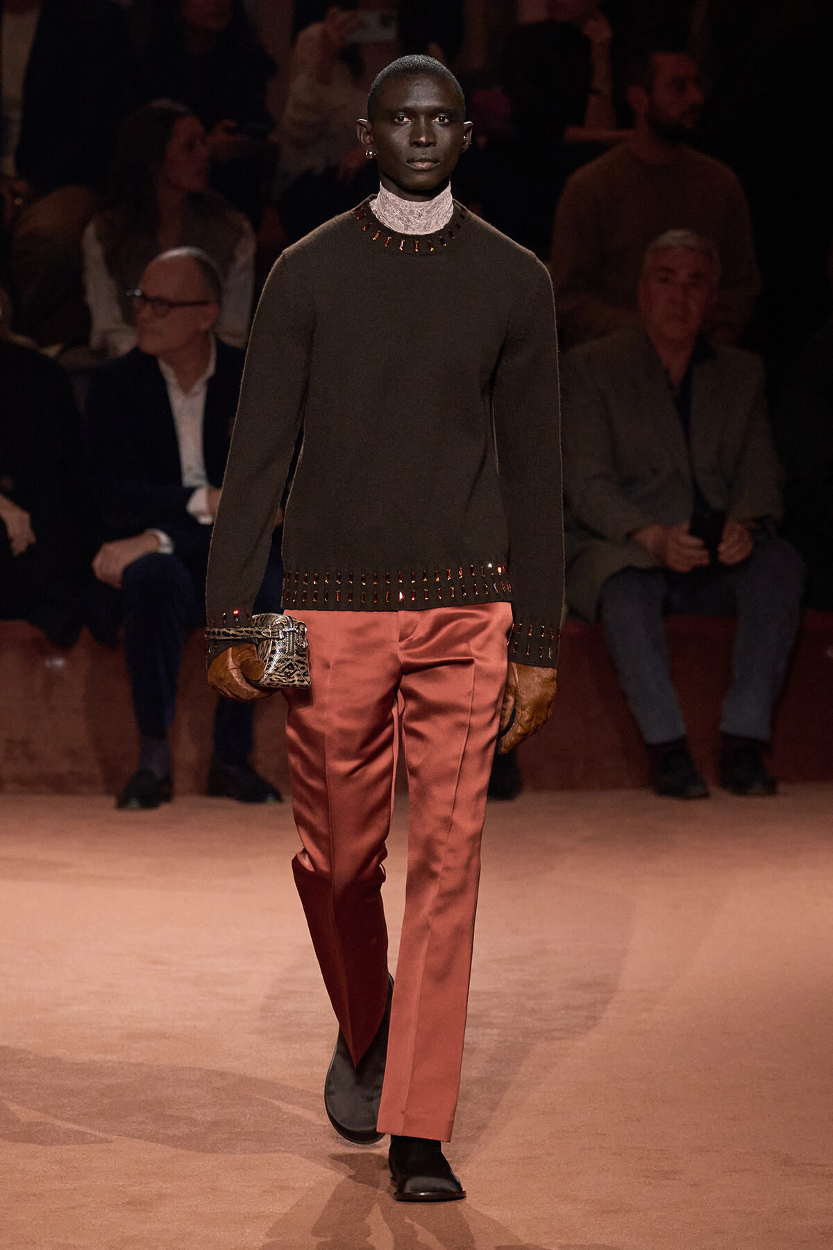 Male model walks a fashion runway in a dark brown studded sweater, pink satin pants, and brown gloves, carrying a patterned clutch.