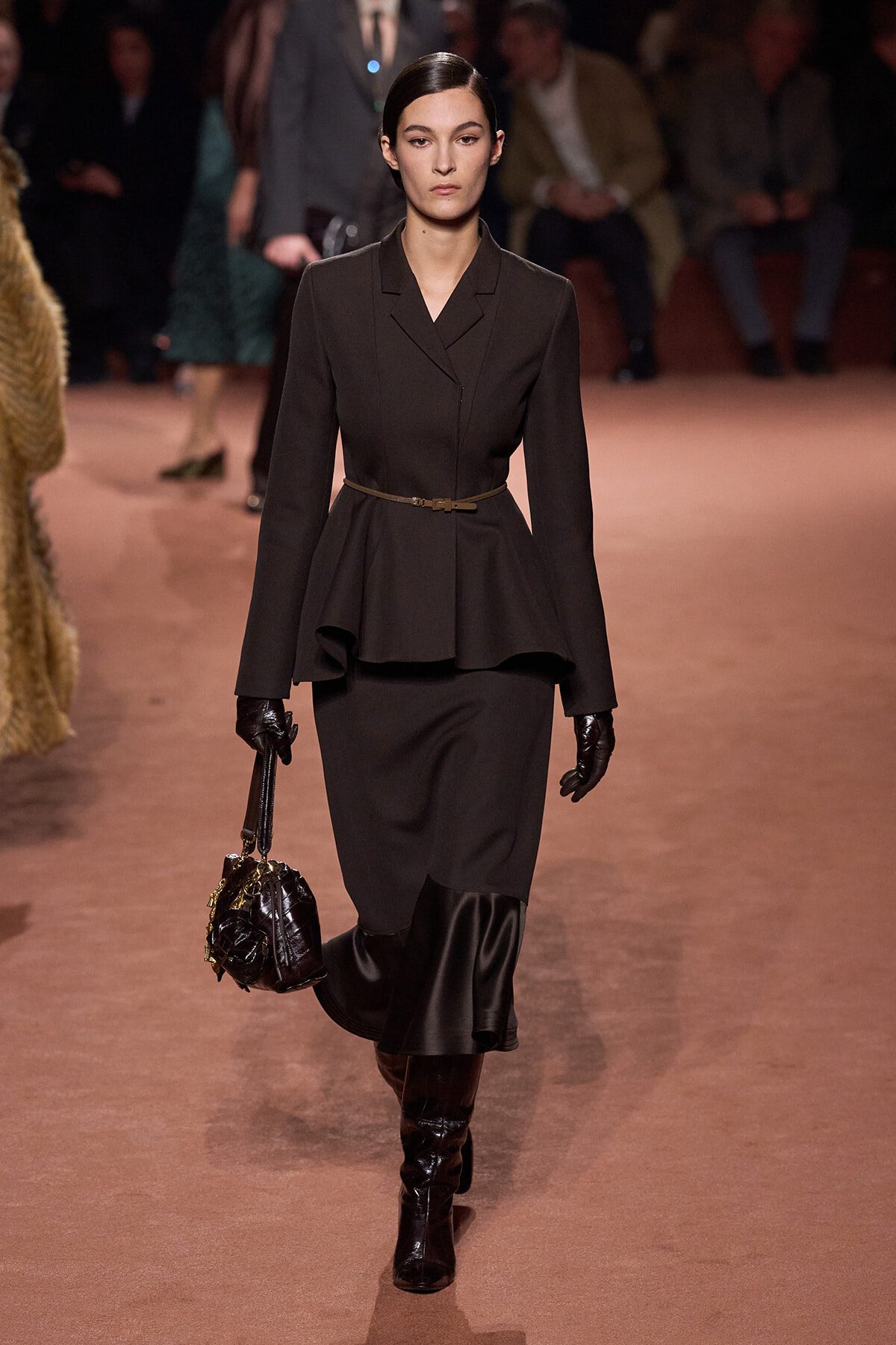 Model walks a dark tailored peplum blazer and matching skirt on a fashion runway, holding a quilted black handbag with gloves and knee boots