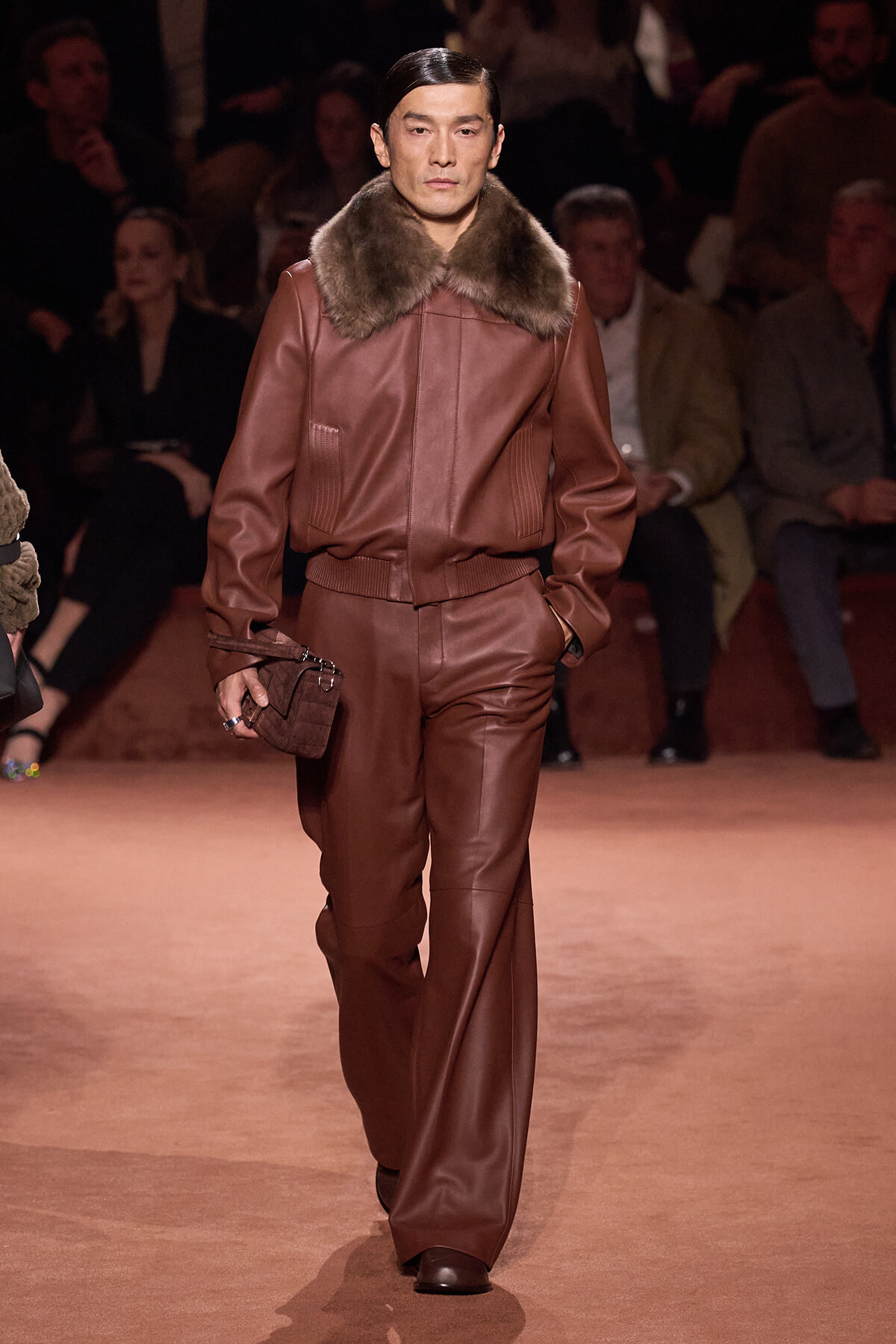 Male model walks a runway in a brown leather suit with a fur collar and matching brown accessories.