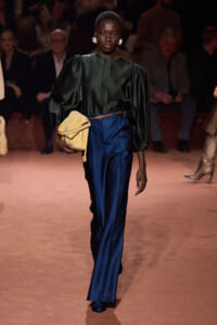 Model walking a runway in a dark green puff-sleeve blouse, cobalt satin pants, tan belt, and a pale yellow clutch with gold earrings nearby.